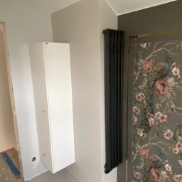 Remontly - Łazienka w trakcie wykańczania z białą szafką wiszącą, czarnym grzejnikiem żeberkowym i płytkami z motywem pawia i kwiatów, widoczne ślady prac remontowych.
