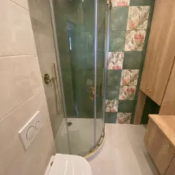 Nowoczesna łazienka z kabiną prysznicową z złotymi detalami, toaletą podwieszaną i zielono-kwiatową mozaiką na ścianie, drewniana szafka z blatem.