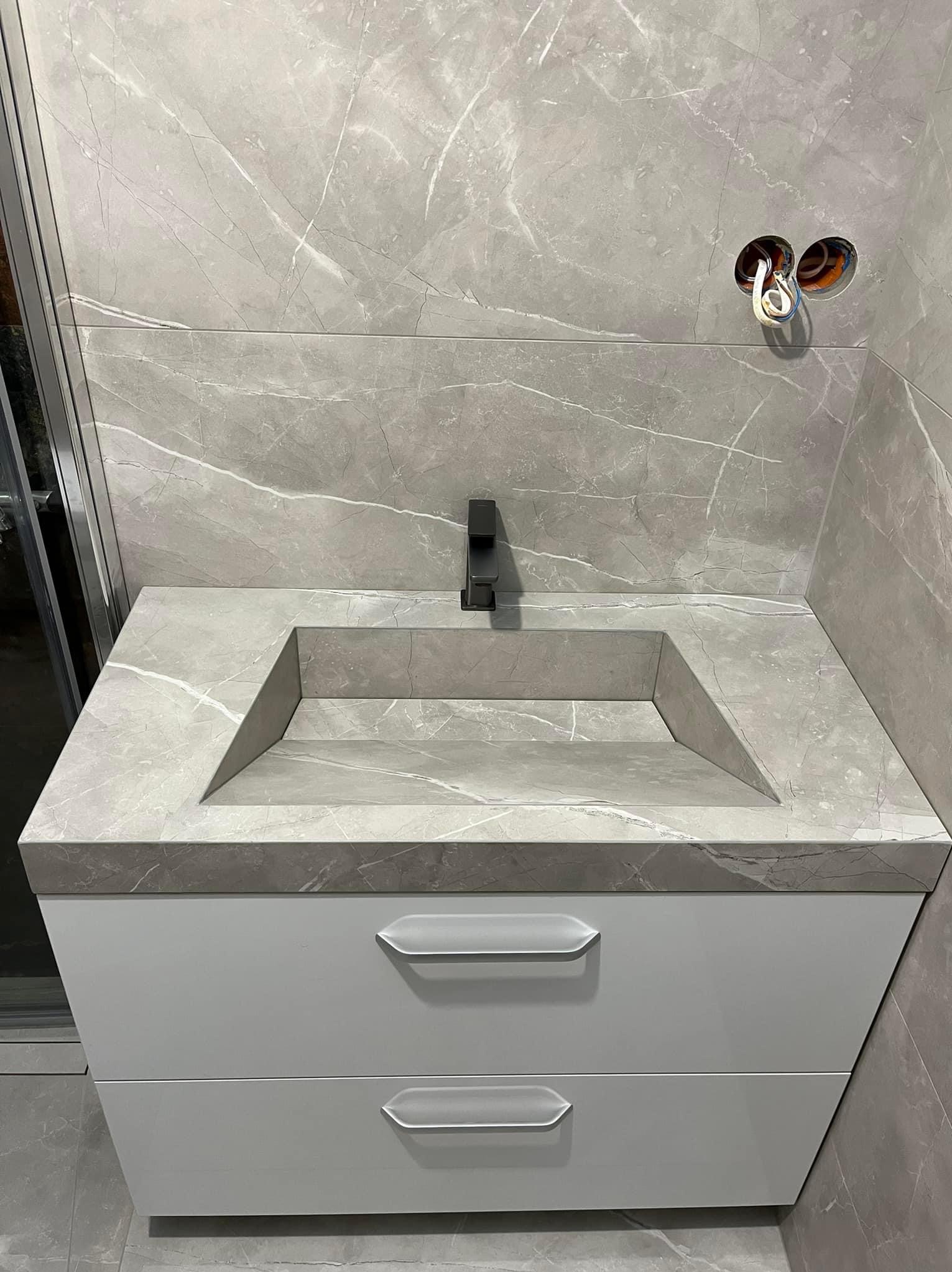 Szara marmurowa umywalka wpuszczona w blat z szafką z dwoma szufladami, nowoczesna bateria w kolorze czarnym, szare marmurowe płytki na ścianie, widoczne otwory z kablami elektrycznymi.