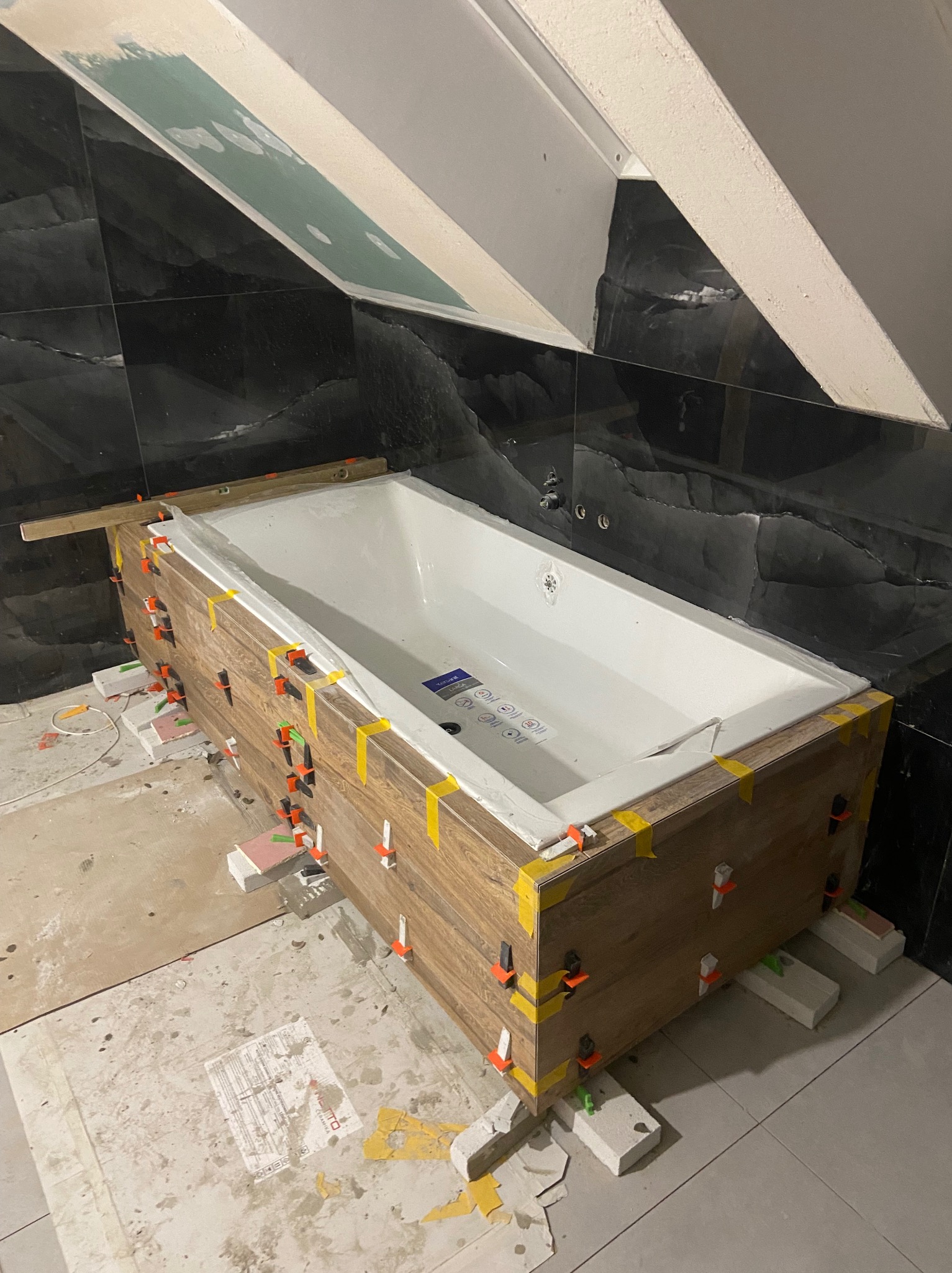 Obudowa wanny z płytek imitujących drewno w trakcie montażu, poziomica na wannie, czarne płytki na ścianach, widoczne kliny i poziomice, poddasze.