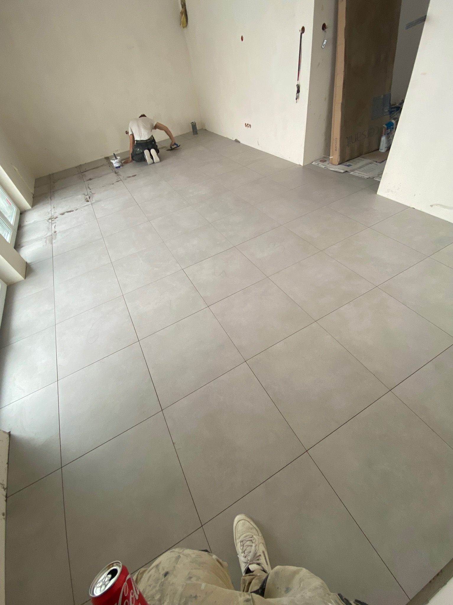 Układanie jasnych, kwadratowych płytek podłogowych w dużym, jasnym pomieszczeniu; widoczny pracownik w oddali, wyrównujący płytki oraz puszka coli i robocze buty w dolnym rogu.