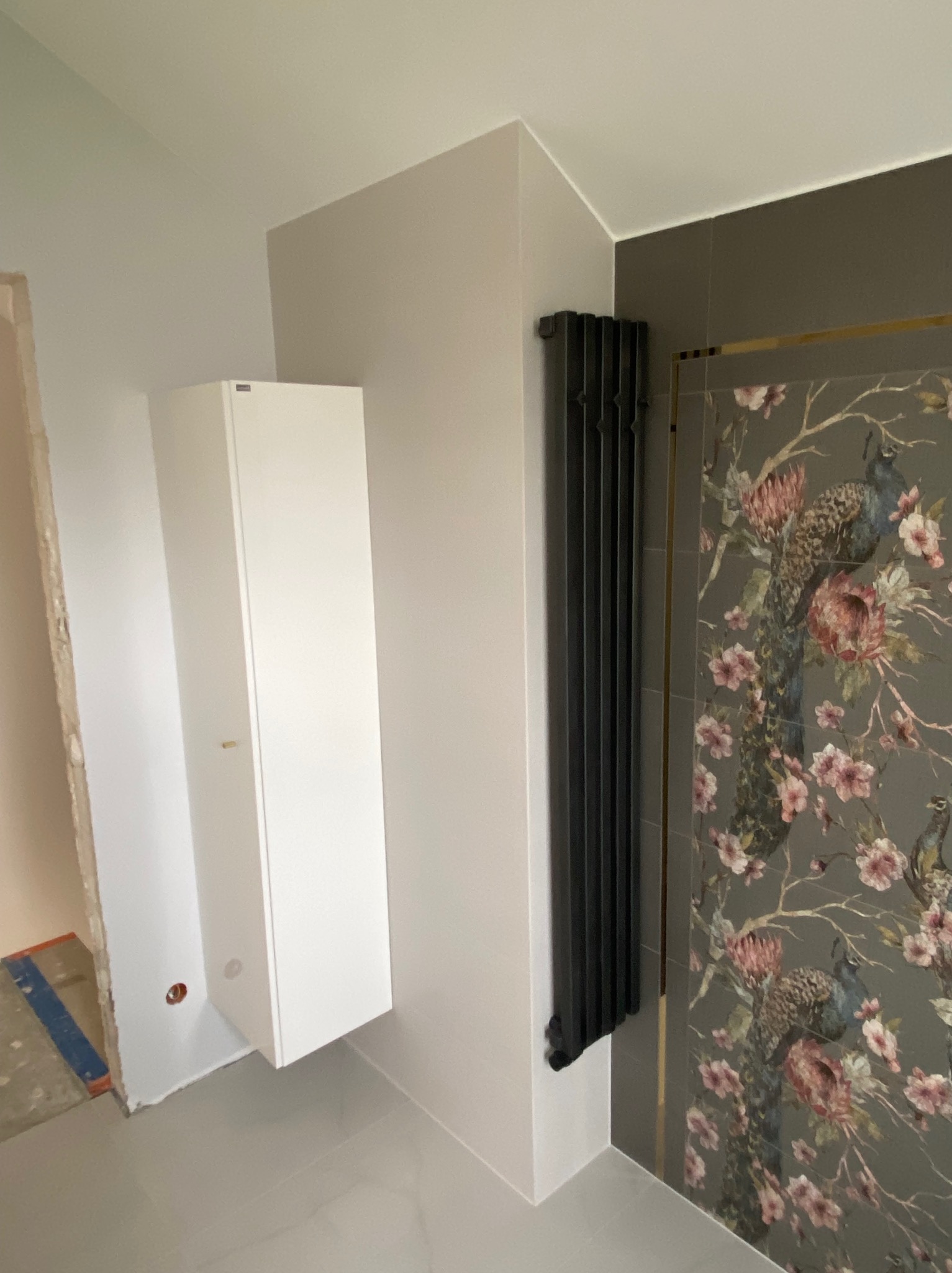 Łazienka w trakcie wykańczania z białą szafką wiszącą, czarnym grzejnikiem żeberkowym i płytkami z motywem pawia i kwiatów, widoczne ślady prac remontowych.