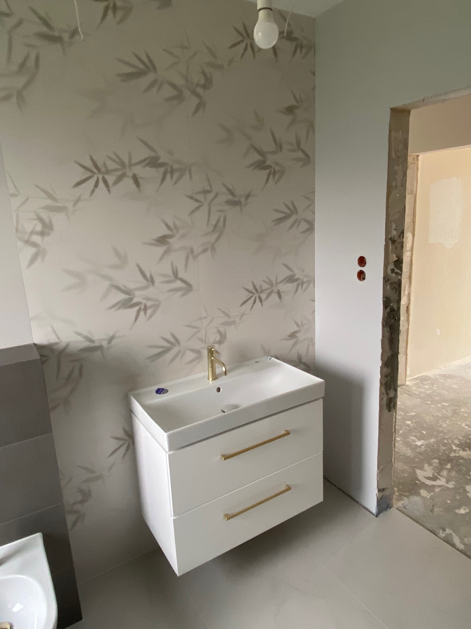 Biała szafka pod umywalkę z złotymi uchwytami, zamontowana na tle ściany z motywem bambusa, widoczny fragment umywalki po lewej stronie oraz surowa ościeżnica drzwi po prawej.