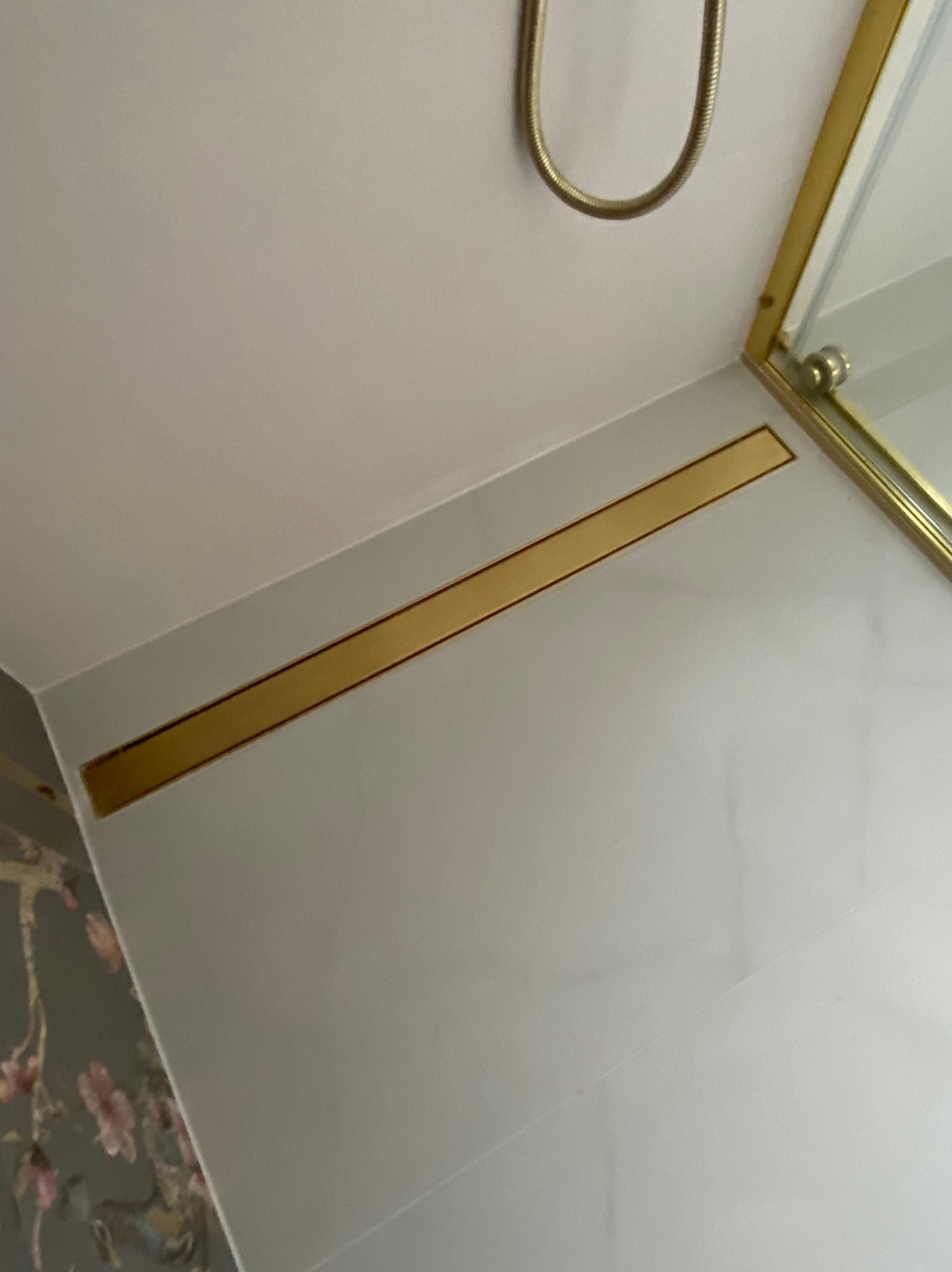 Wykończona łazienka z białymi płytkami i dekoracyjnym złotym pasem pod sufitem oraz fragmentem lustra w złotej ramie i słuchawką prysznicową.