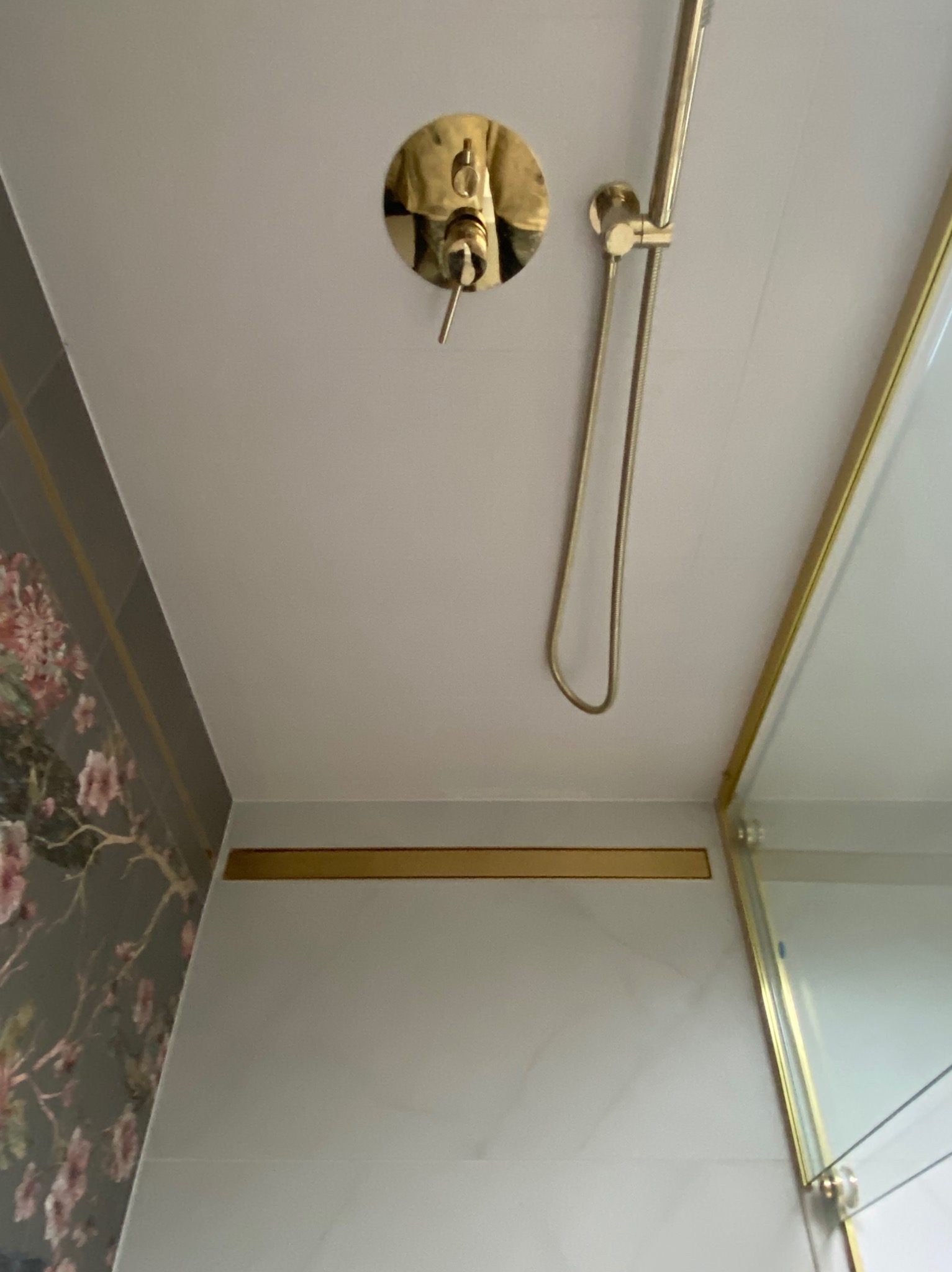 Detal wykończenia łazienki: złota armatura prysznicowa, w tym bateria i deszczownica, na tle białych płytek oraz fragment ściany z tapetą w kwiatowy wzór.