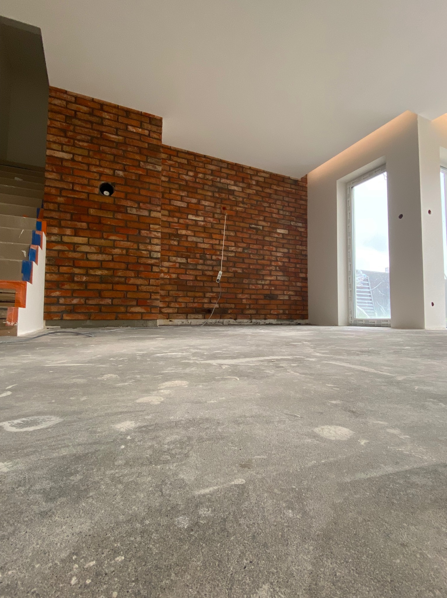 Surowy pokój z ceglaną ścianą, widoczne przewody elektryczne, szary beton na podłodze, okno z białymi ramami, schody w tle z zabezpieczeniami w kolorze pomarańczowym i niebieskim.