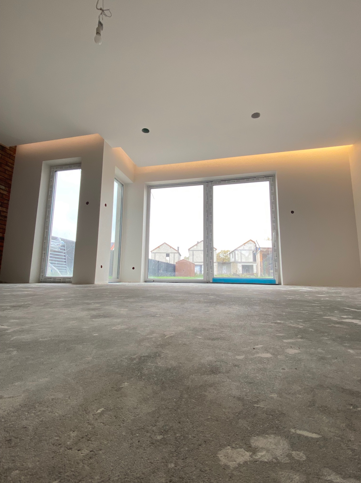 Surowy betonowy podkład w jasnym pokoju z oknami tarasowymi i oświetleniem LED podsufitowym, widoczne ślady po pracach budowlanych.