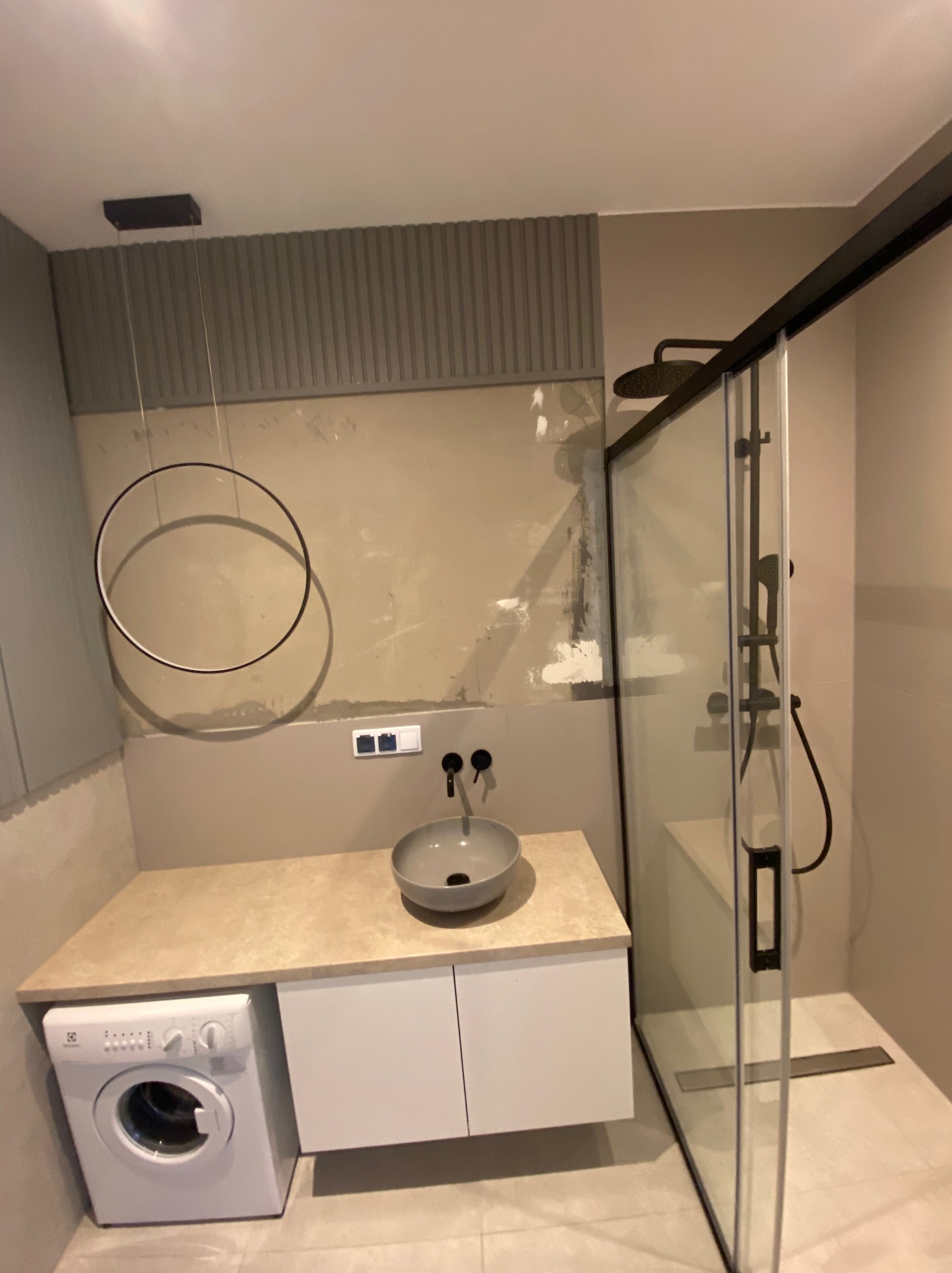 Wnętrze łazienki w trakcie prac wykończeniowych, widoczne ślady po gładzi na ścianach, nowoczesna kabina prysznicowa z czarnymi elementami, okrągła lampa wisząca oraz pralka pod umywalką.