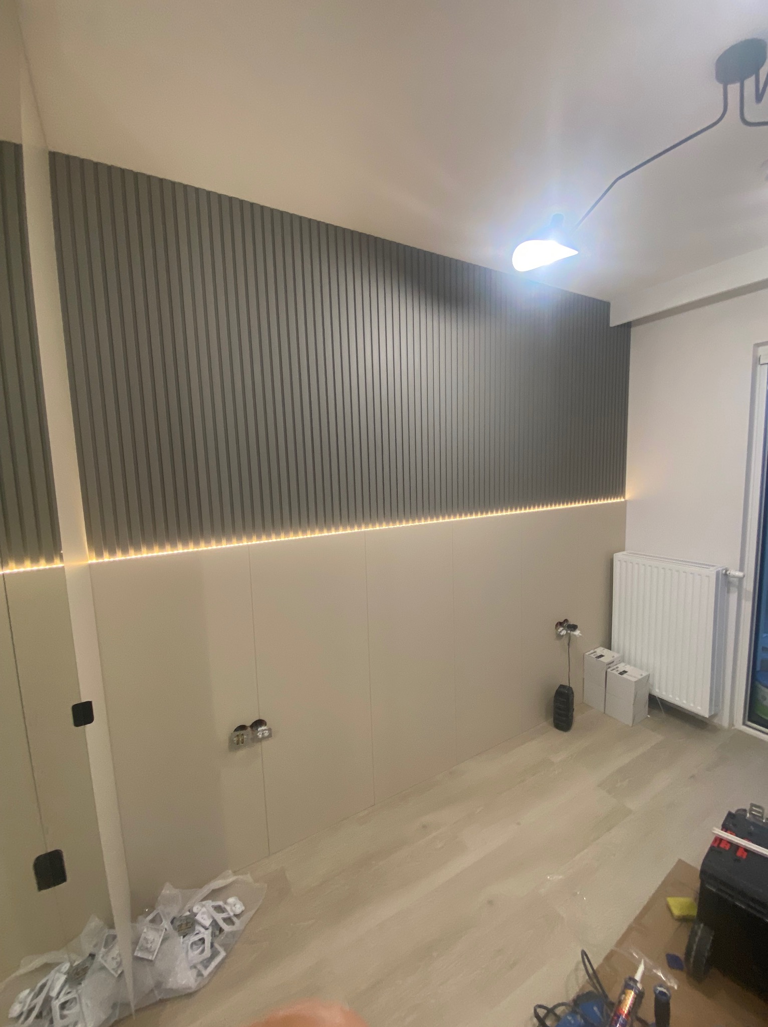 Pokój w trakcie prac, ściana wykończona panelami pionowymi w odcieniach szarości z podświetleniem LED, widoczne puszki elektryczne, grzejnik, narzędzia i materiały budowlane.