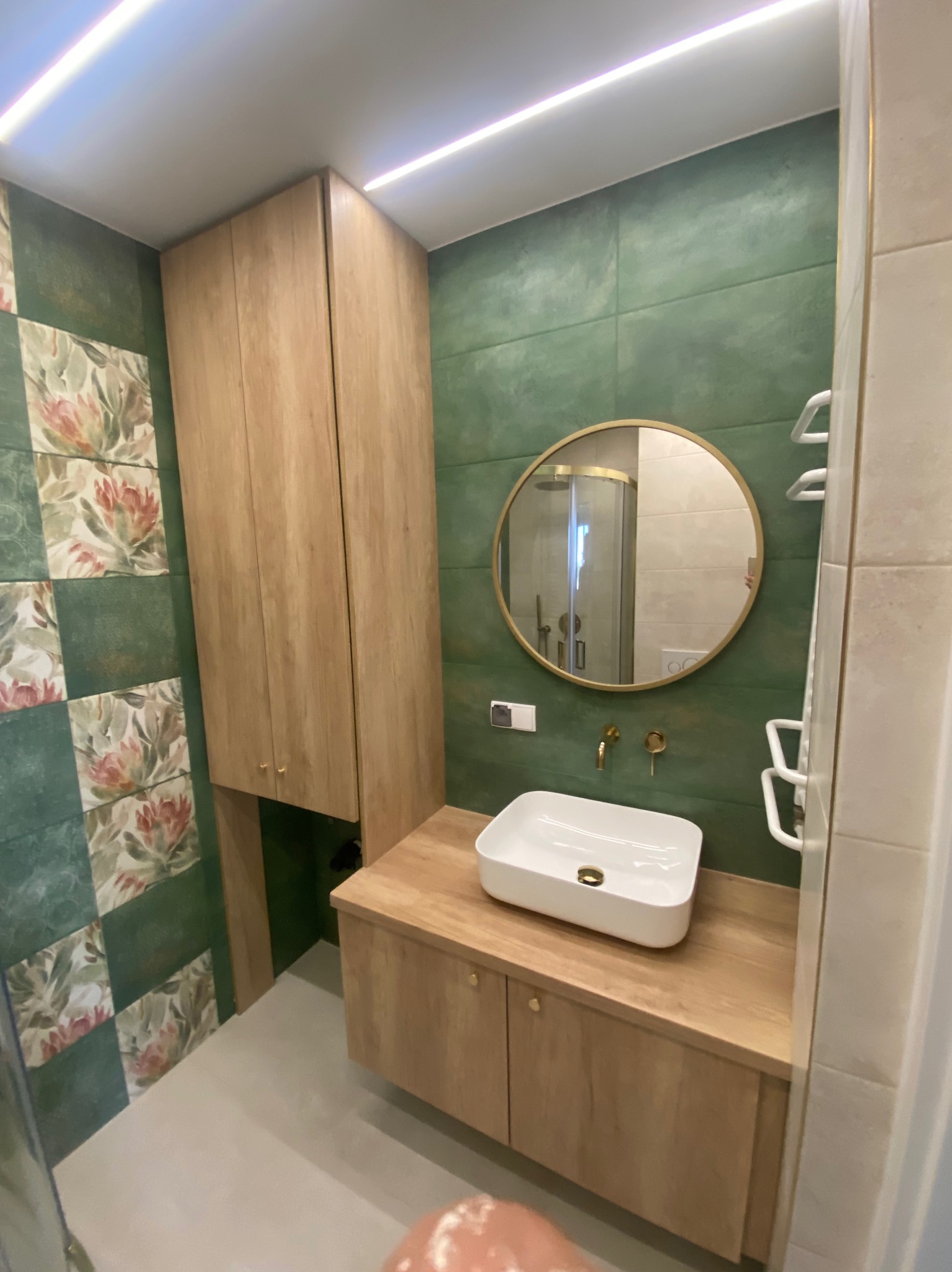 Nowoczesna łazienka z zielonymi płytkami, drewnianą szafką pod umywalką i okrągłym lustrem ze złotą ramą, widok na kabinę prysznicową w odbiciu lustra.