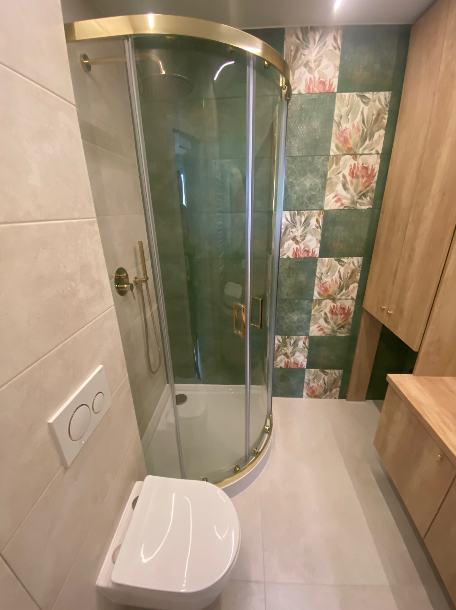 Nowoczesna łazienka z kabiną prysznicową z złotymi detalami, toaletą podwieszaną i zielono-kwiatową mozaiką na ścianie, drewniana szafka z blatem.