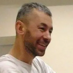 Uśmiechnięty mężczyzna z krótkimi, siwiejącymi włosami patrzy w prawo, widoczny od szyi w górę.