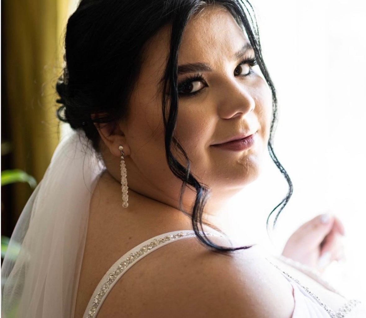 Panna młoda z ciemnymi włosami i subtelnym makijażem oczu, spoglądająca przez ramię, z widocznymi długimi kolczykami i welonem.