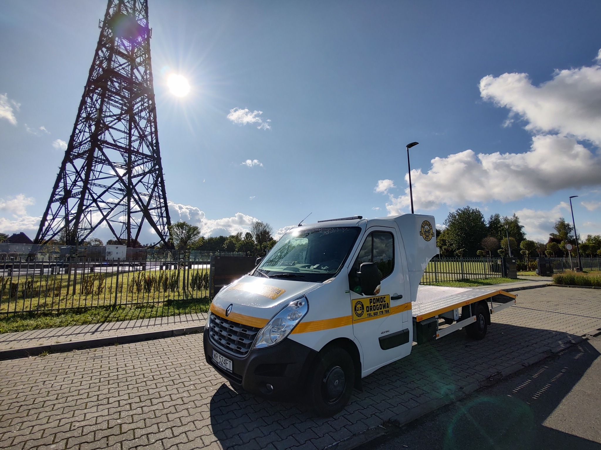 Biała laweta z żółtymi pasami i logiem 'Pomoc Drogowa' zaparkowana na brukowanej powierzchni, z wysoką wieżą radiową w tle i jasnym słońcem na błękitnym niebie z chmurami.