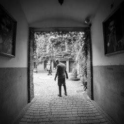 Kamerą i aparatem - Czarno-białe zdjęcie przedstawia mężczyznę idącego przez kamienny dziedziniec w kierunku budynku porośniętego bluszczem, z portretami w ramkach po bokach.