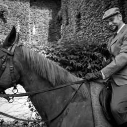 Kamerą i aparatem - Czarno-białe zdjęcie eleganckiego mężczyzny w tweedowym garniturze i kaszkiecie siedzącego na koniu, trzymającego fajkę w skórzanej rękawiczce, z bluszczem porastającym budynek w tle.