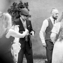 Kamerą i aparatem - Czarno-białe zdjęcie grupy osób w eleganckich strojach w stylu retro, prawdopodobnie podczas przygotowań do uroczystości, z mężczyzną w kaszkiecie sprawdzającym coś na smartfonie.