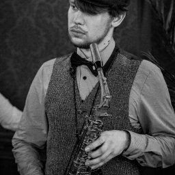 Kamerą i aparatem - Czarno-białe zdjęcie mężczyzny w tweedowej kamizelce i kaszkiecie, trzymającego saksofon, z delikatnym uśmiechem i widocznym tatuażem na szyi.