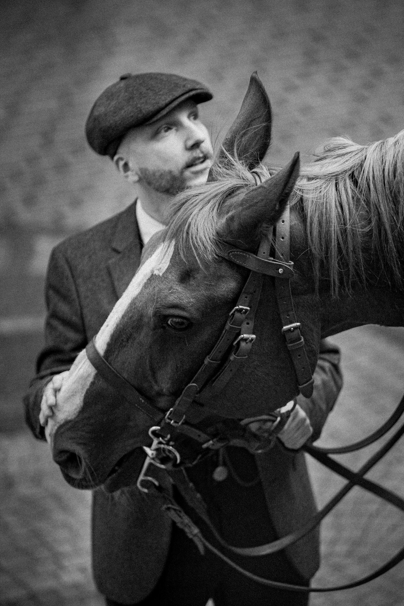 Czarno-białe zdjęcie mężczyzny w tweedowej marynarce i kaszkiecie, obejmującego głowę konia z białą strzałką na pysku i skórzanym ogłowiem.