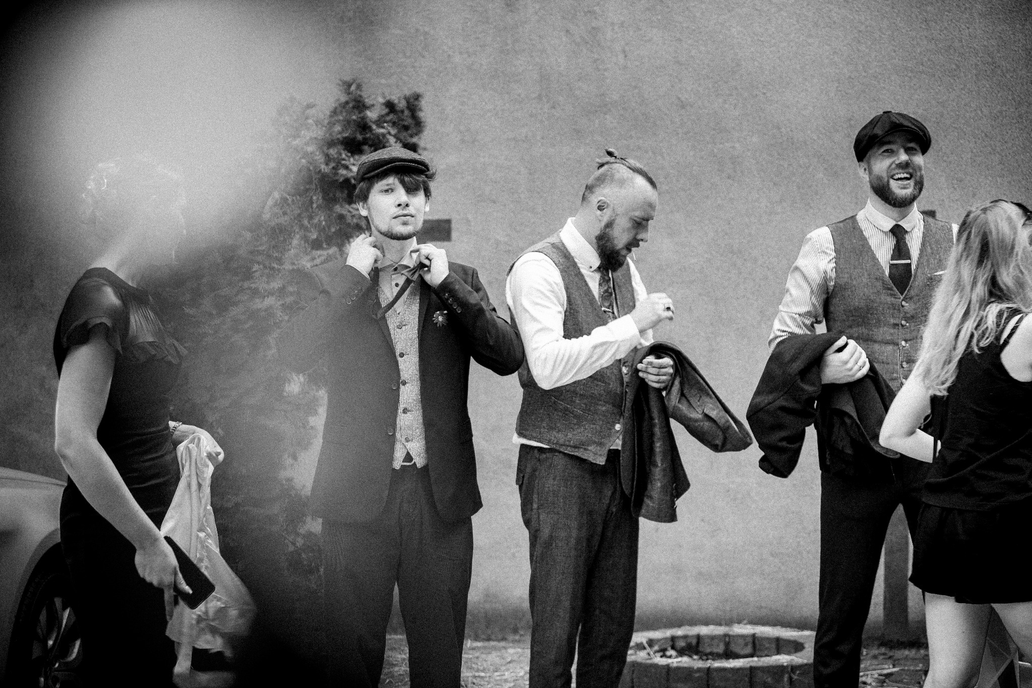 Czarno-białe zdjęcie grupy osób w eleganckich strojach w stylu retro: mężczyźni w tweedowych kamizelkach i kaszkietach, jeden poprawia krawat, drugi trzyma marynarkę, trzeci się śmieje; kobiety...