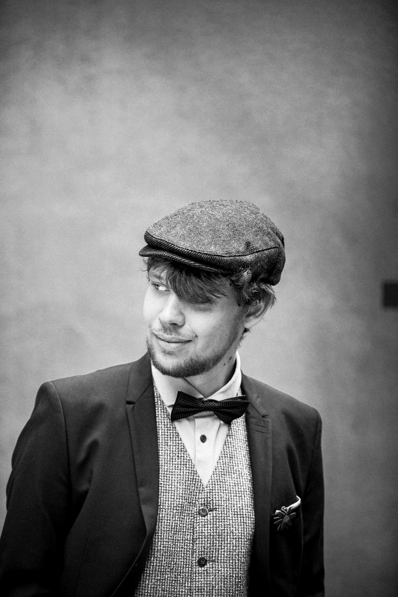 Portret mężczyzny w tweedowej czapce i muszce, ubrany w garnitur i kamizelkę, spoglądającego w bok z lekkim uśmiechem, wykonany w czerni i bieli.