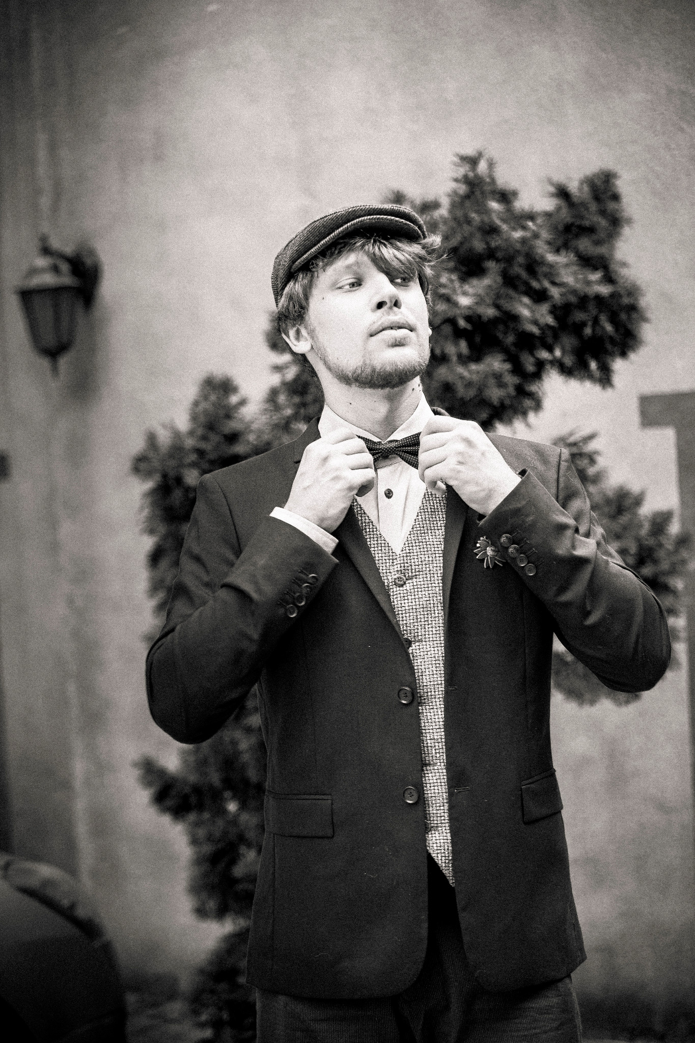 Mężczyzna w tweedowej czapce i garniturze poprawia muchę na tle ściany i krzewu, zdjęcie czarno-białe.