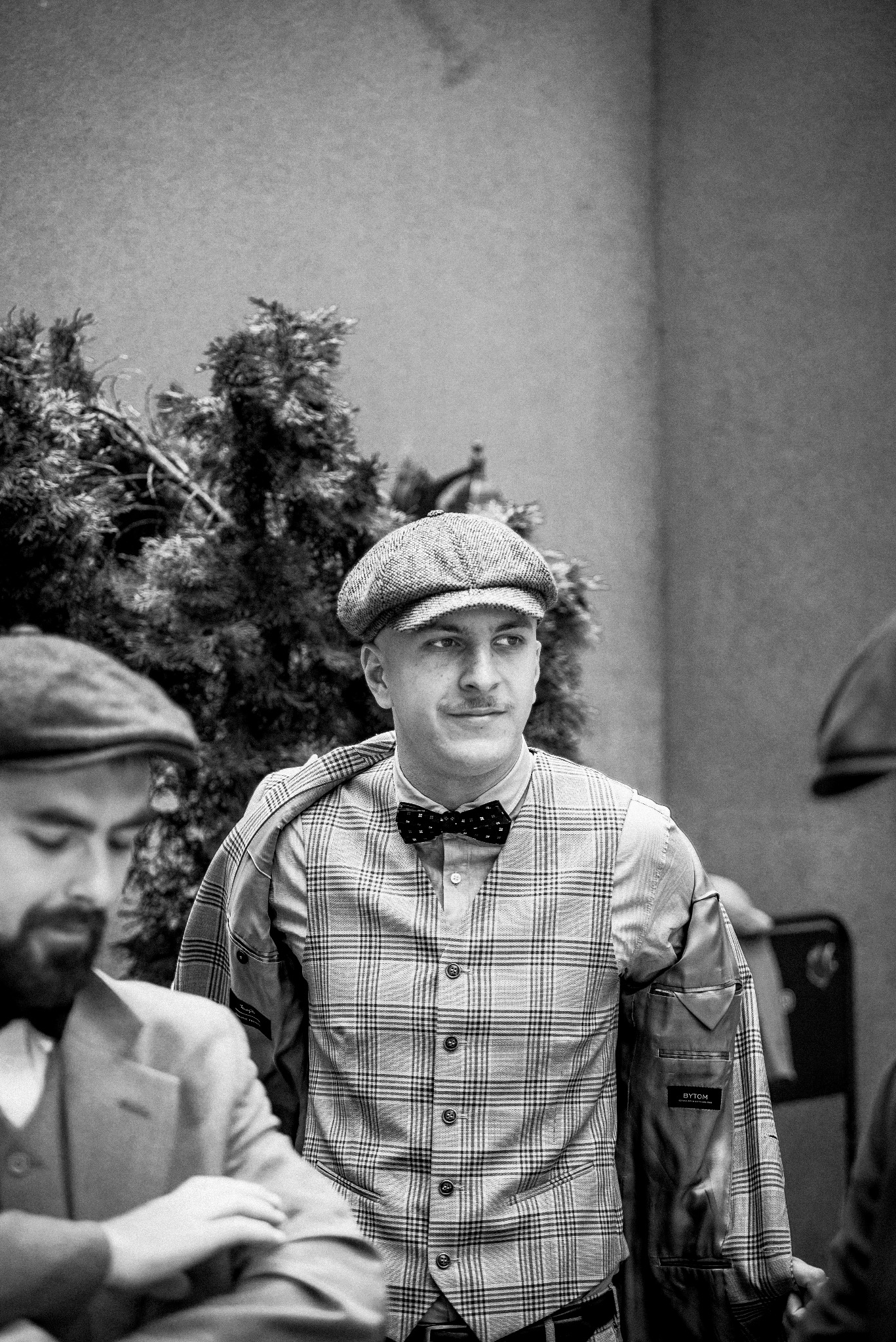 Mężczyzna z wąsami w kraciastej kamizelce, muszce i kaszkiecie, z marynarką przerzuconą przez ramię, w towarzystwie innych osób w retro strojach, na neutralnym tle.