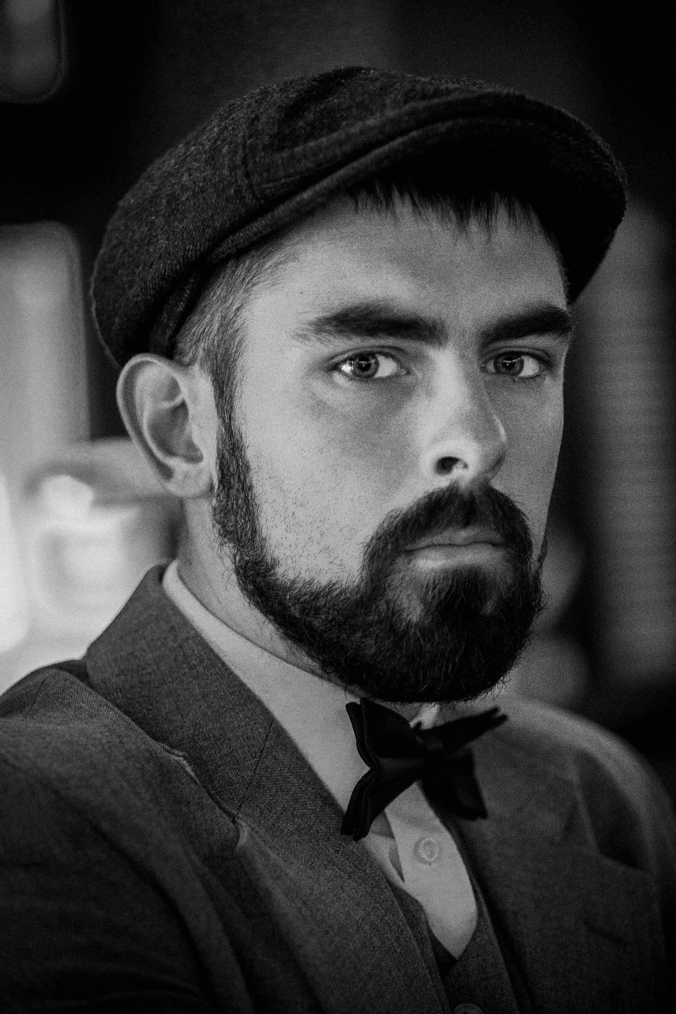 Czarno-biały portret mężczyzny z brodą w tweedowej czapce i muszce, patrzącego prosto w obiektyw.