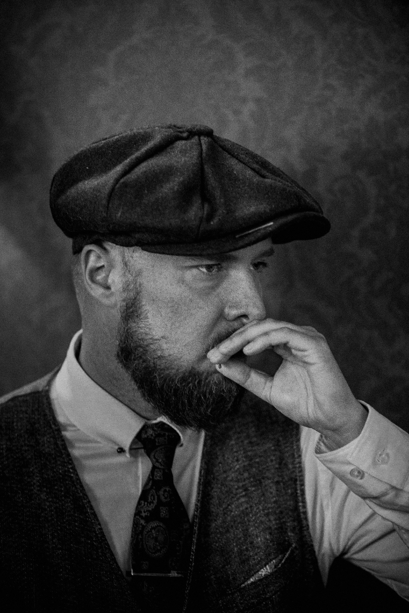 Czarno-białe portretowe ujęcie brodatego mężczyzny w kaszkiecie i kamizelce, z papierosem w dłoni, w stylu retro, wykonane w Krakowie.