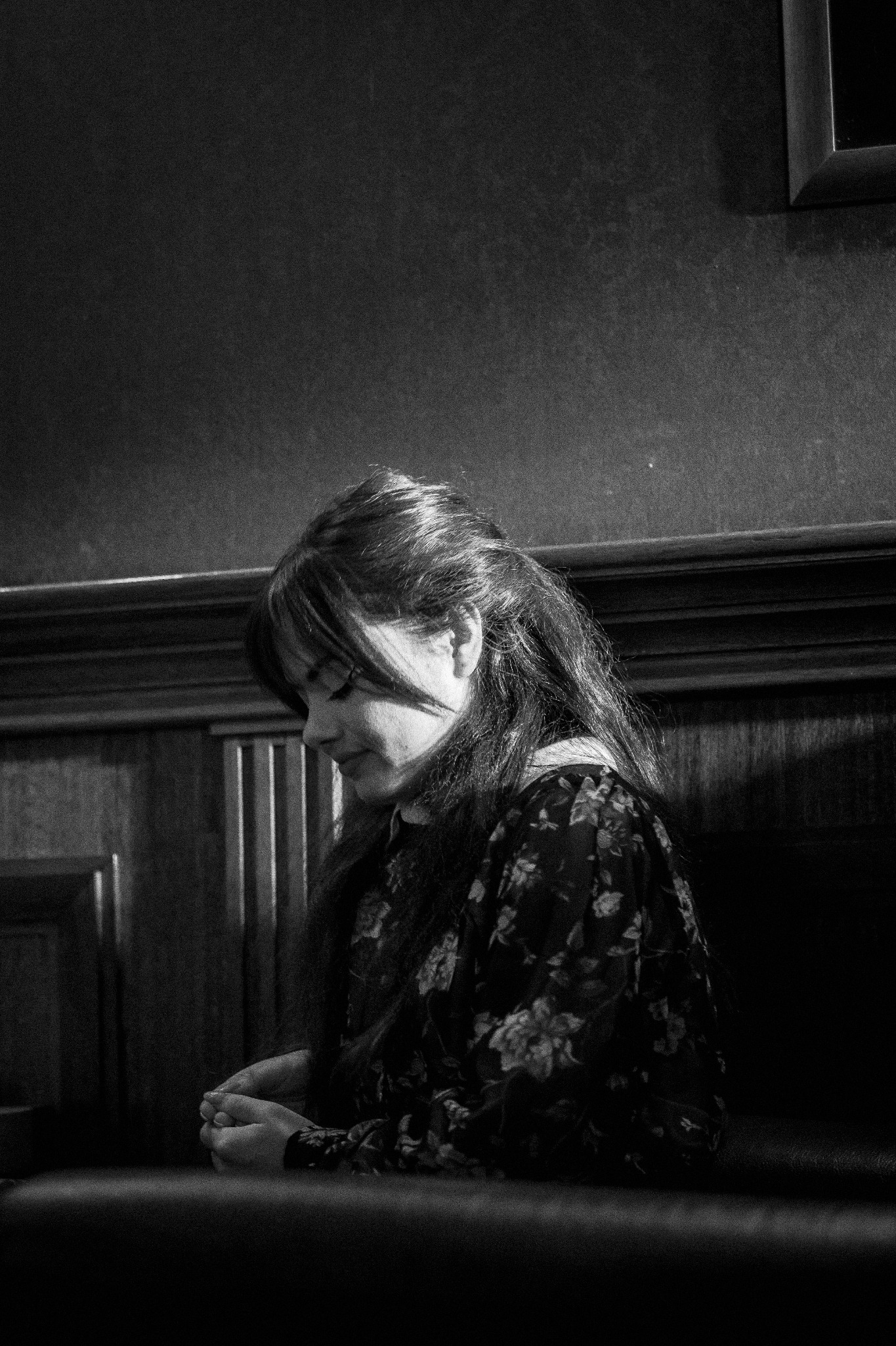 Portret kobiety z profilu w czarno-białej tonacji, siedzącej na tle drewnianej boazerii i ciemnej ściany, z rękami splecionymi w kolanach i wzrokiem skierowanym w dół.
