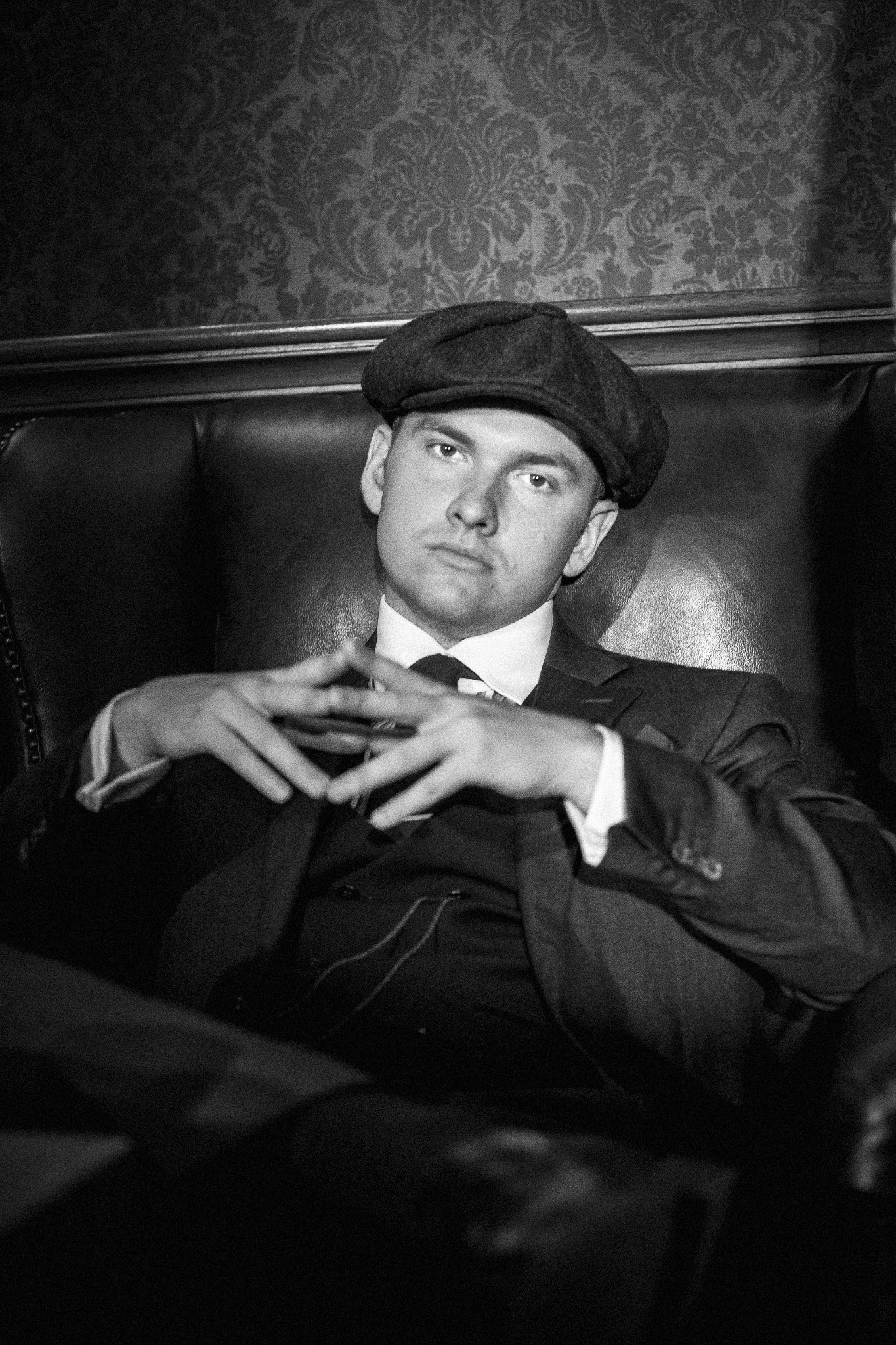 Czarno-białe zdjęcie mężczyzny w garniturze i kaszkiecie, siedzącego w skórzanym fotelu z rękami splecionymi przed sobą na tle wzorzystej tapety.