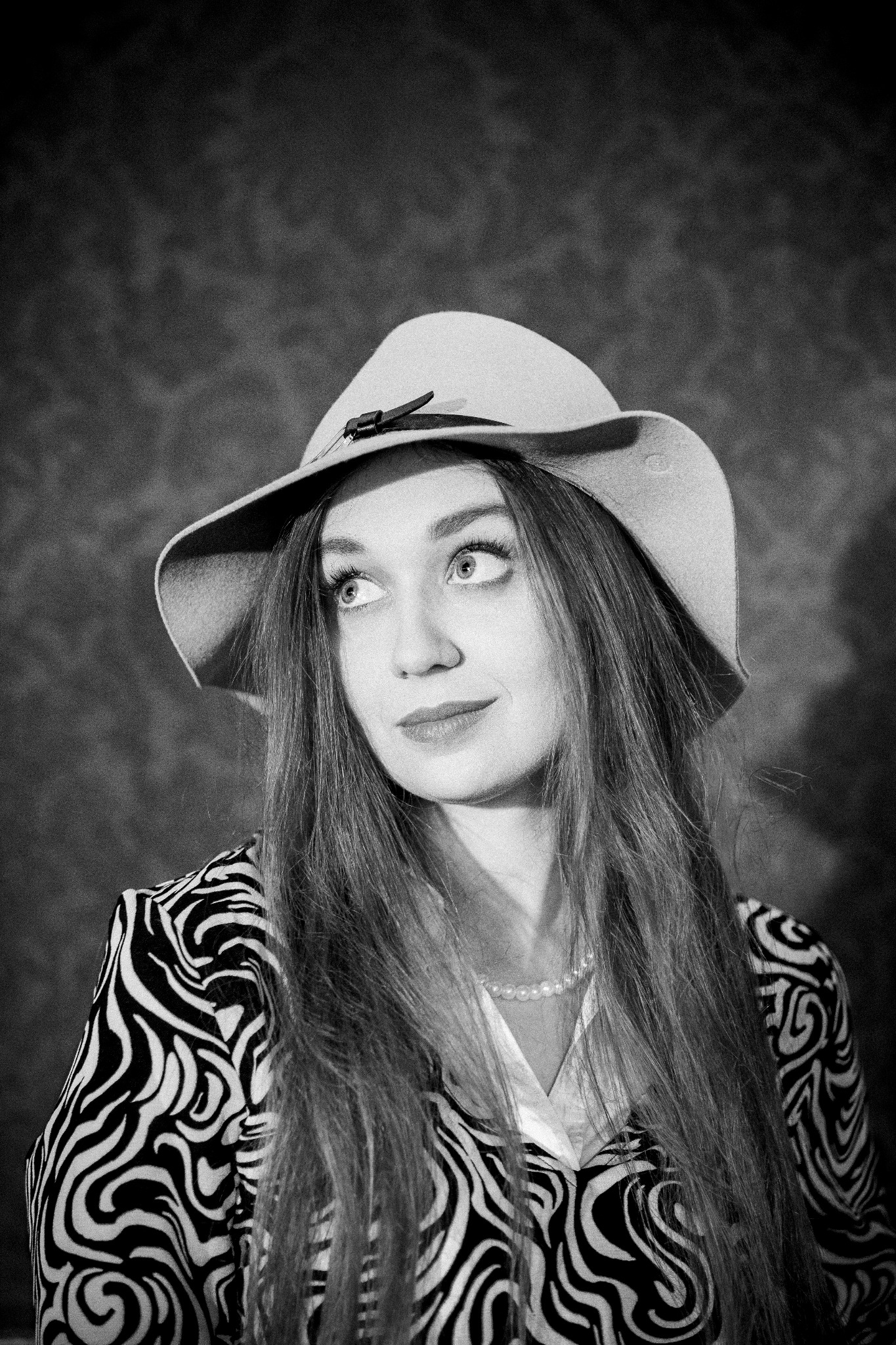 Portret kobiety w kapeluszu z szerokim rondem i bluzce w zebrę, spoglądającej w górę w czarno-białej tonacji, z perłowym naszyjnikiem.
