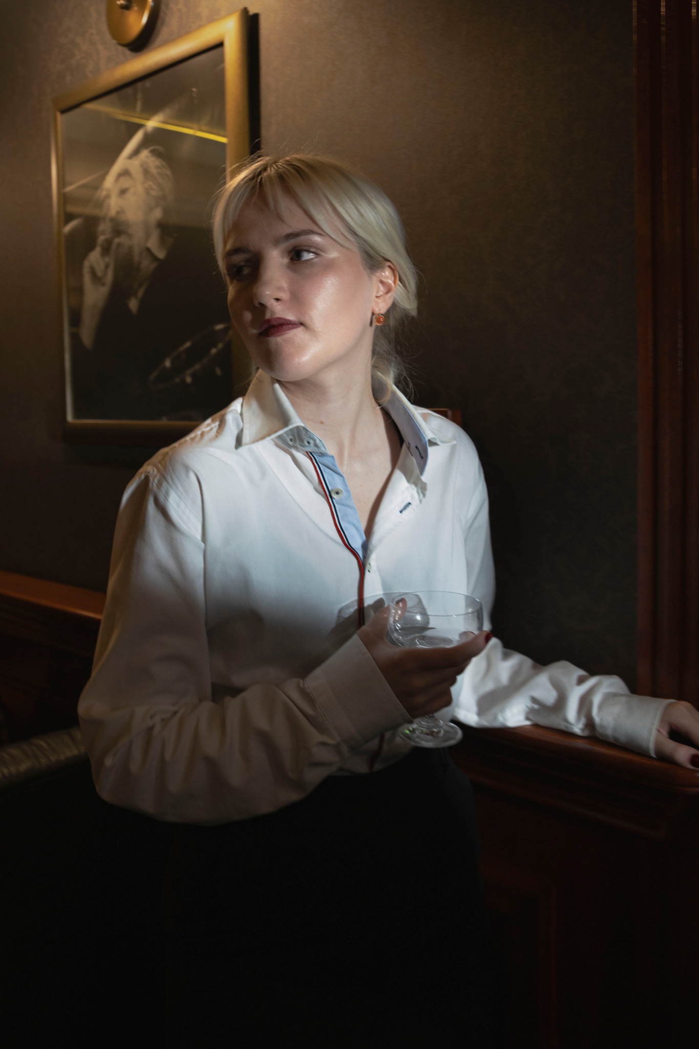 Portret kobiety w białej koszuli, trzymającej pusty kieliszek na tle ściany z obrazem w złotej ramie.