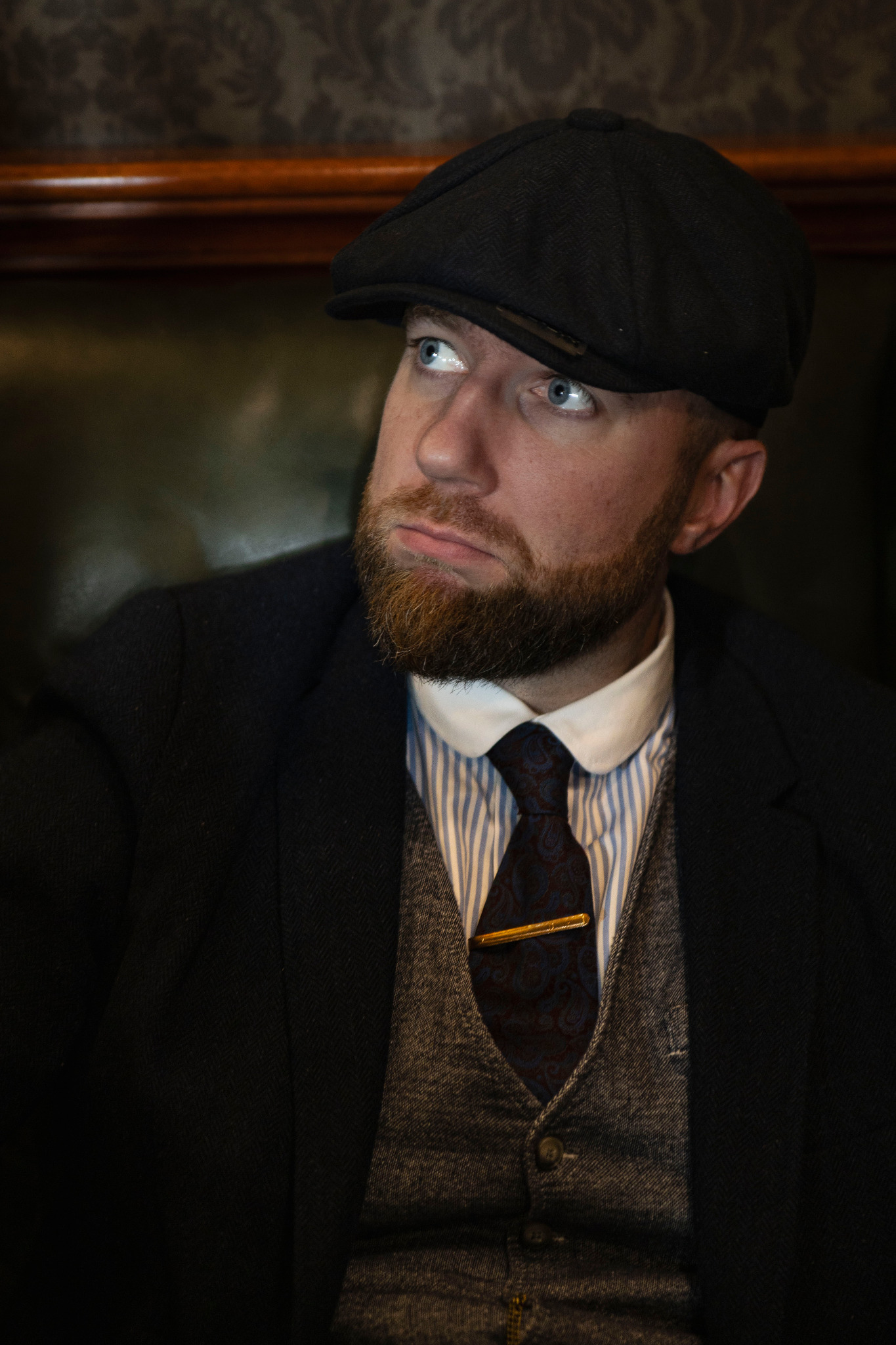 Portret mężczyzny z brodą w tweedowej czapce, kamizelce i marynarce, patrzącego w górę w stylu retro.