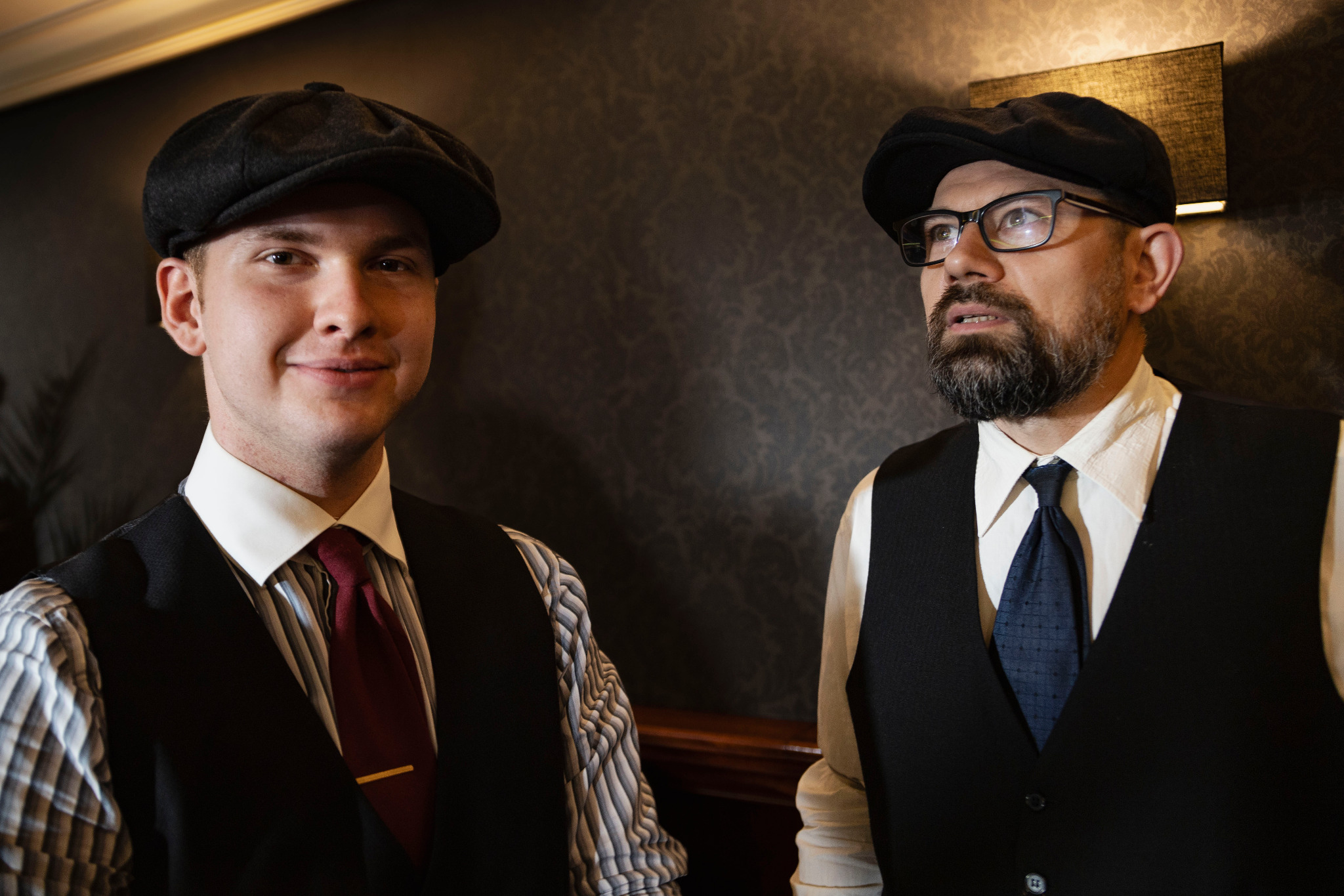 Portret dwóch mężczyzn w czarnych kamizelkach i kaszkietach, jeden z uśmiechem, drugi w okularach, na tle ciemnej ściany z lampą.