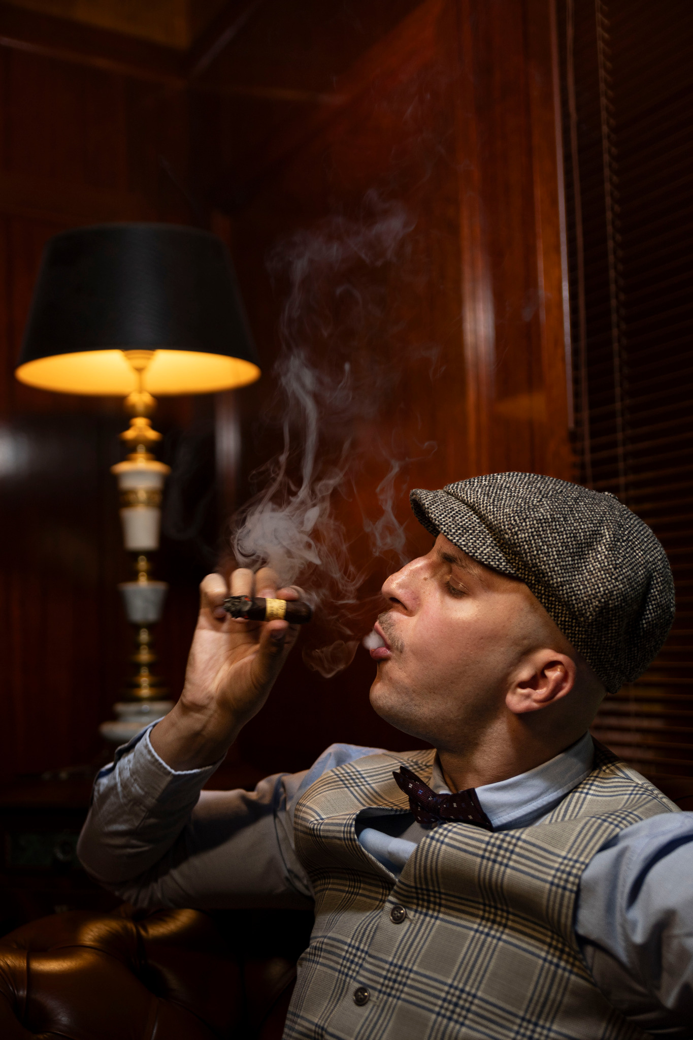 Mężczyzna w kaszkiecie i kamizelce w kratę, palący cygaro w dymie, oświetlony ciepłym światłem lampy na tle ciemnego, drewnianego wnętrza.