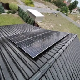 Panele słoneczne na błyszczącym, czarnym dachu w Osjakowie, widok z góry, w tle zabudowania i zieleń.