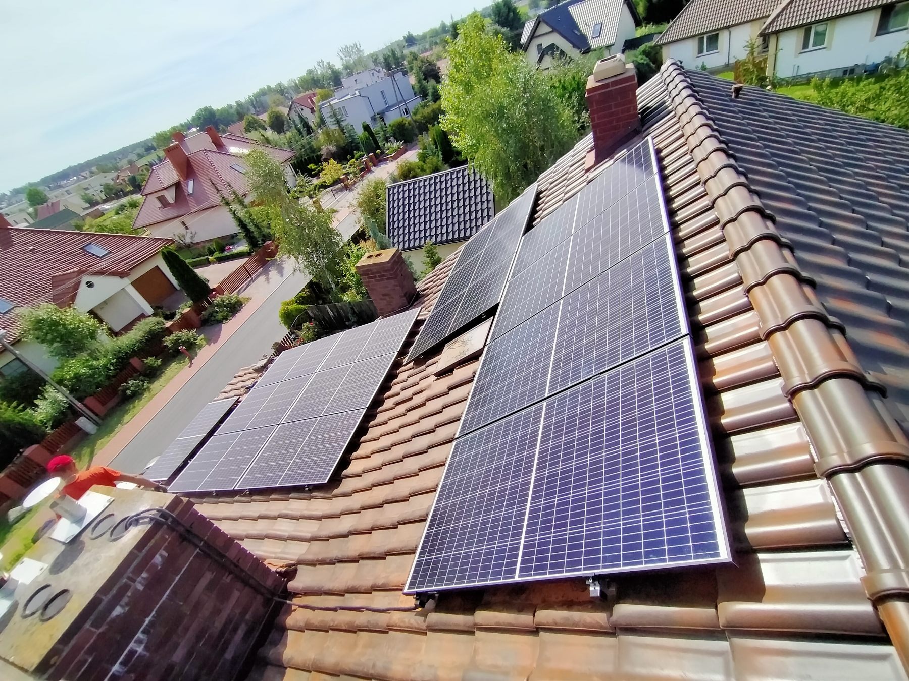 Panele słoneczne zamontowane na brązowym dachu domu jednorodzinnego z widokiem na okoliczne budynki i zieleń w słoneczny dzień.