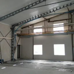 Wnętrze hali przemysłowej w trakcie prac wykończeniowych; widoczne stalowe konstrukcje nośne, drabiny, instalacje pod sufitem i materiały budowlane na podłodze zabezpieczonej folią.