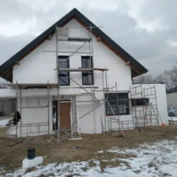 Dom jednorodzinny w trakcie prac ociepleniowych, widoczne rusztowania i styropianowe płyty elewacyjne, częściowo pokryte śniegiem.