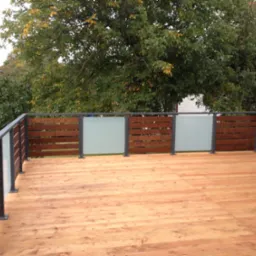 Nowoczesny balkon z drewnianą podłogą i balustradą z poziomych drewnianych paneli oraz matowego szkła, na tle drzew z żółknącymi liśćmi.