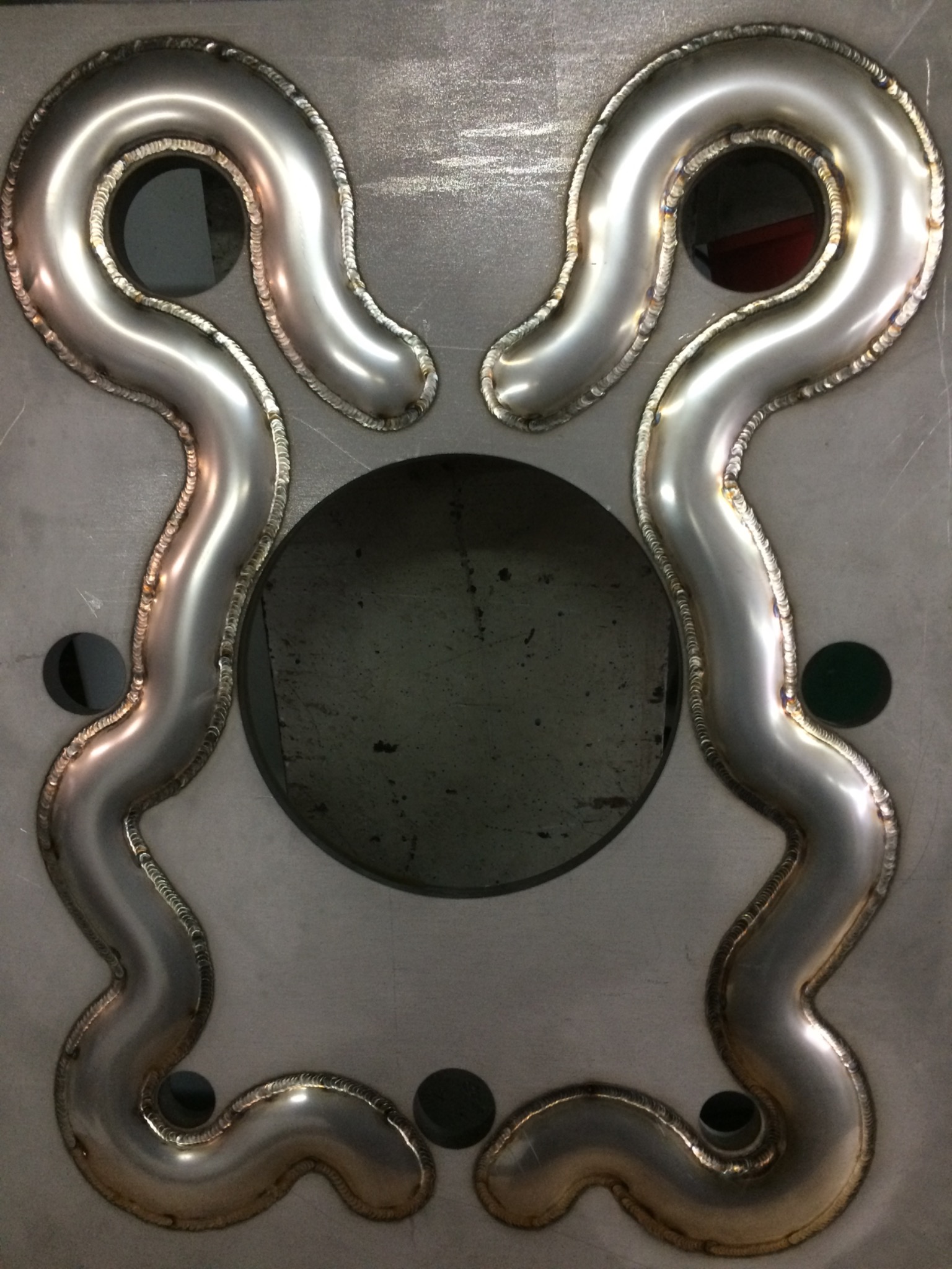 Metalowa konstrukcja z symetrycznie ułożonymi, giętymi rurami, spawana na szarej płycie z widocznymi otworami.