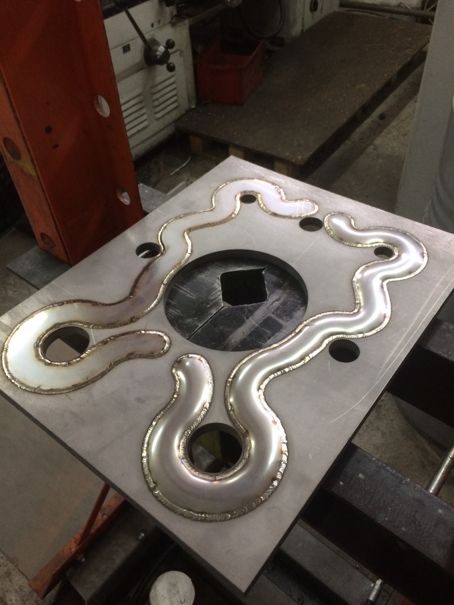 Metalowa płyta z nieregularnym, wypukłym wzorem spawanym na jej powierzchni, widoczne ślady obróbki i otwory.