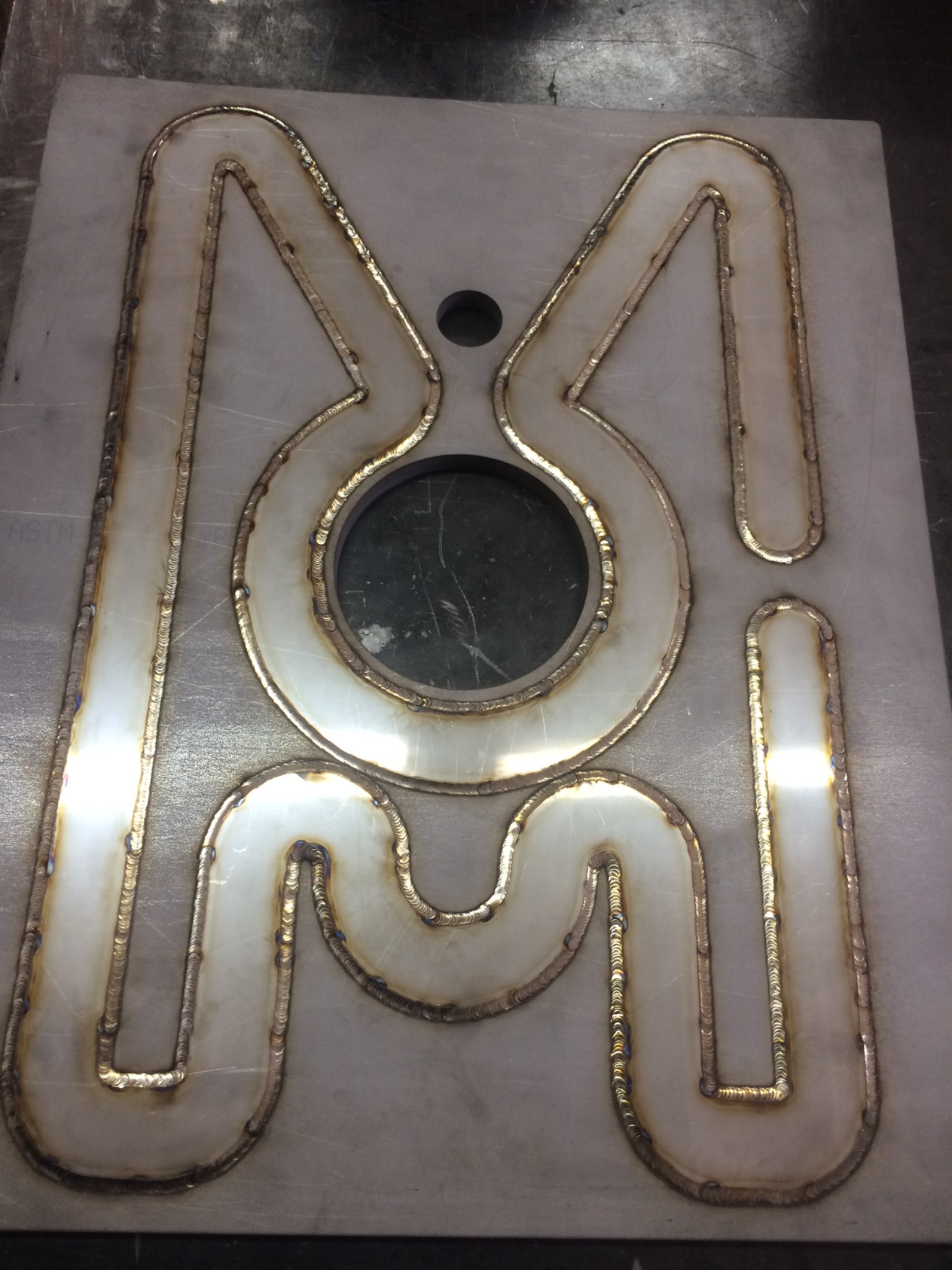 Wypalony laserowo w metalu napis 'MOI' z widocznymi śladami spawania, otworem w literze 'O' i oznaczeniem 'ASTM' na powierzchni.