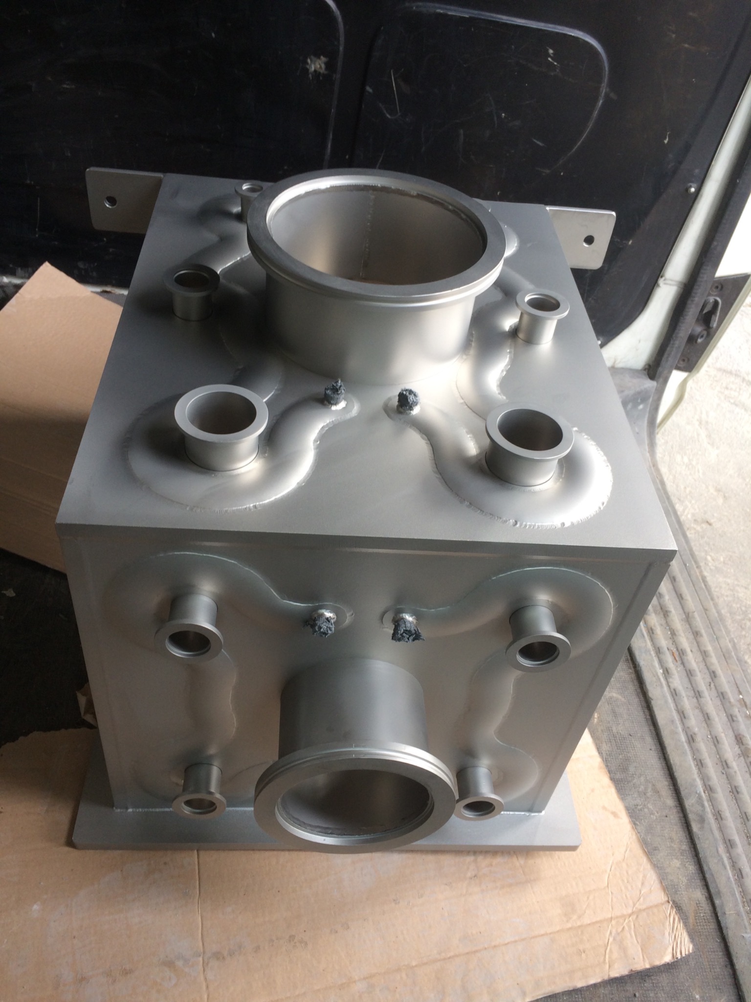 Szczegółowe ujęcie srebrnej, metalowej skrzynki z przyspawanymi rurami o różnych średnicach, widoczne ślady spawania, umieszczona na kartonie wewnątrz pojazdu.