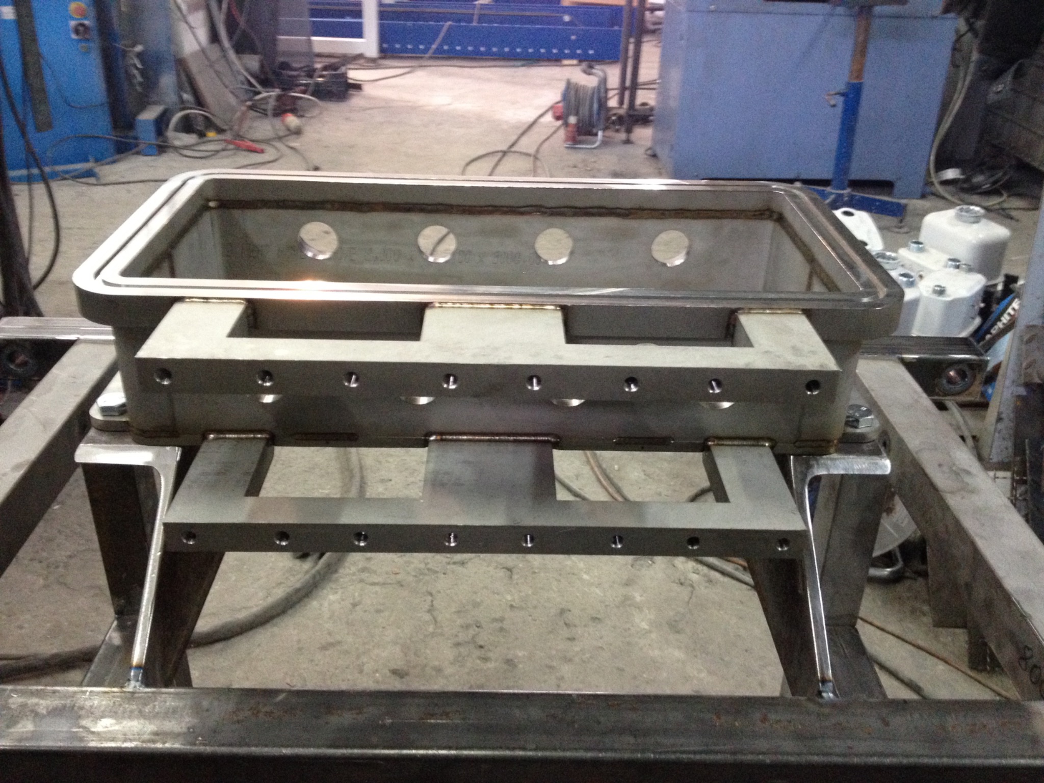 Metalowa konstrukcja skrzynkowa z otworami, wsparta na stalowej ramie, widoczne spawy i elementy montażowe w warsztatowym otoczeniu.