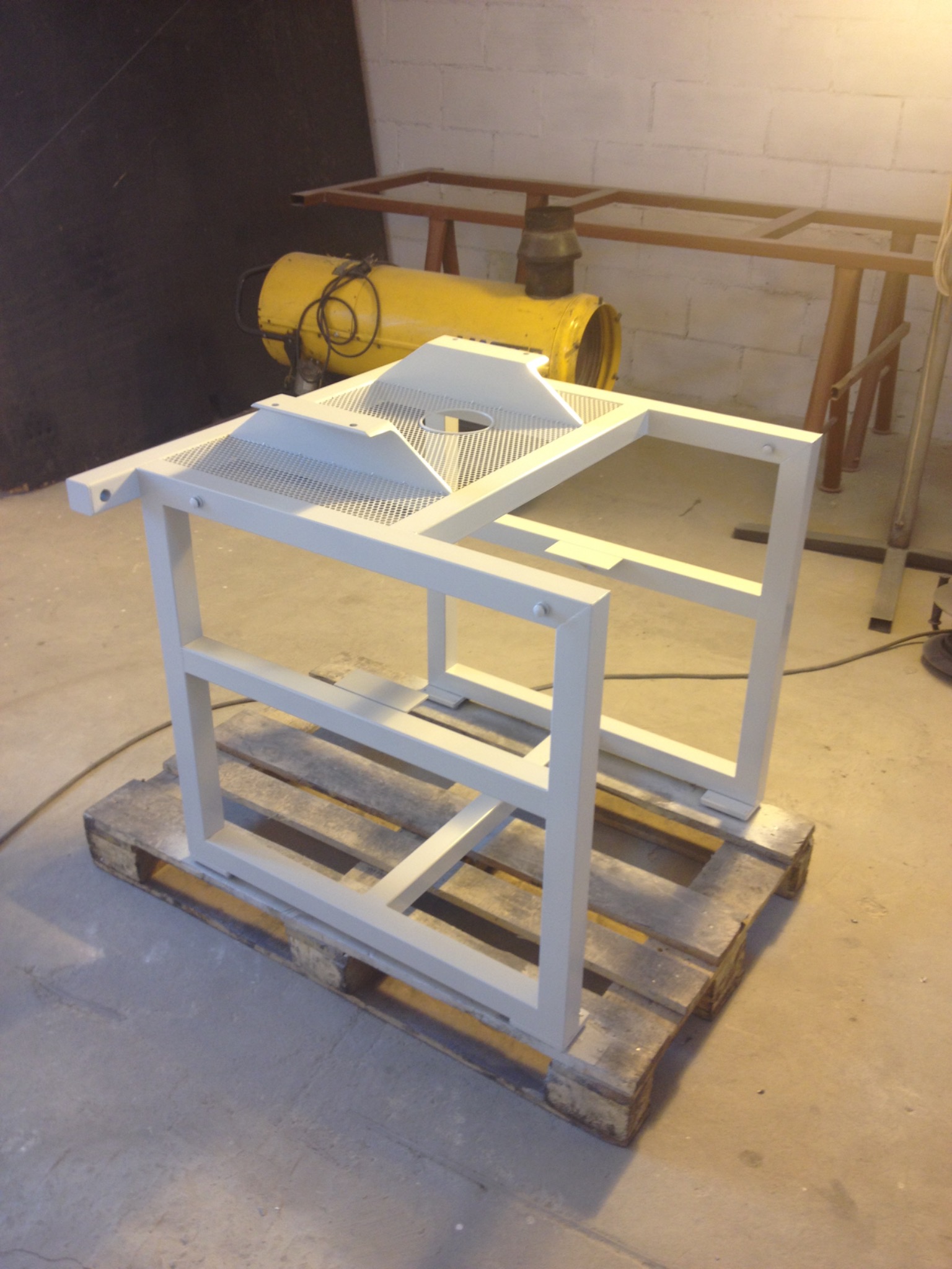 Biała, pomalowana proszkowo, metalowa konstrukcja z perforowanym blatem, stojąca na drewnianej palecie w warsztacie.