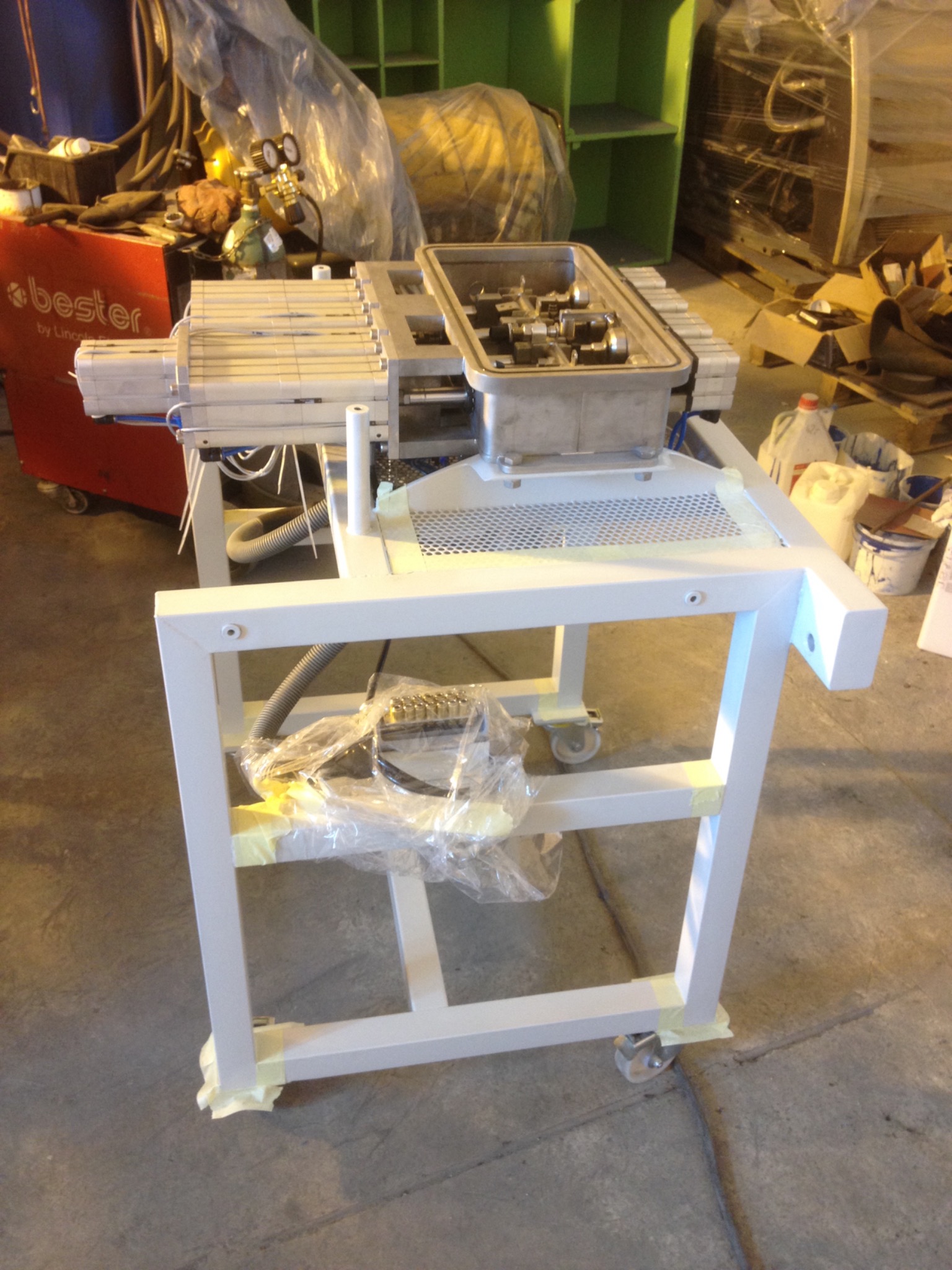 Biała, metalowa konstrukcja na kółkach z elementami zabezpieczonymi taśmą malarską, prawdopodobnie w trakcie malowania lub po nim, z widocznymi elementami hydraulicznymi i pneumatycznymi, na tle...