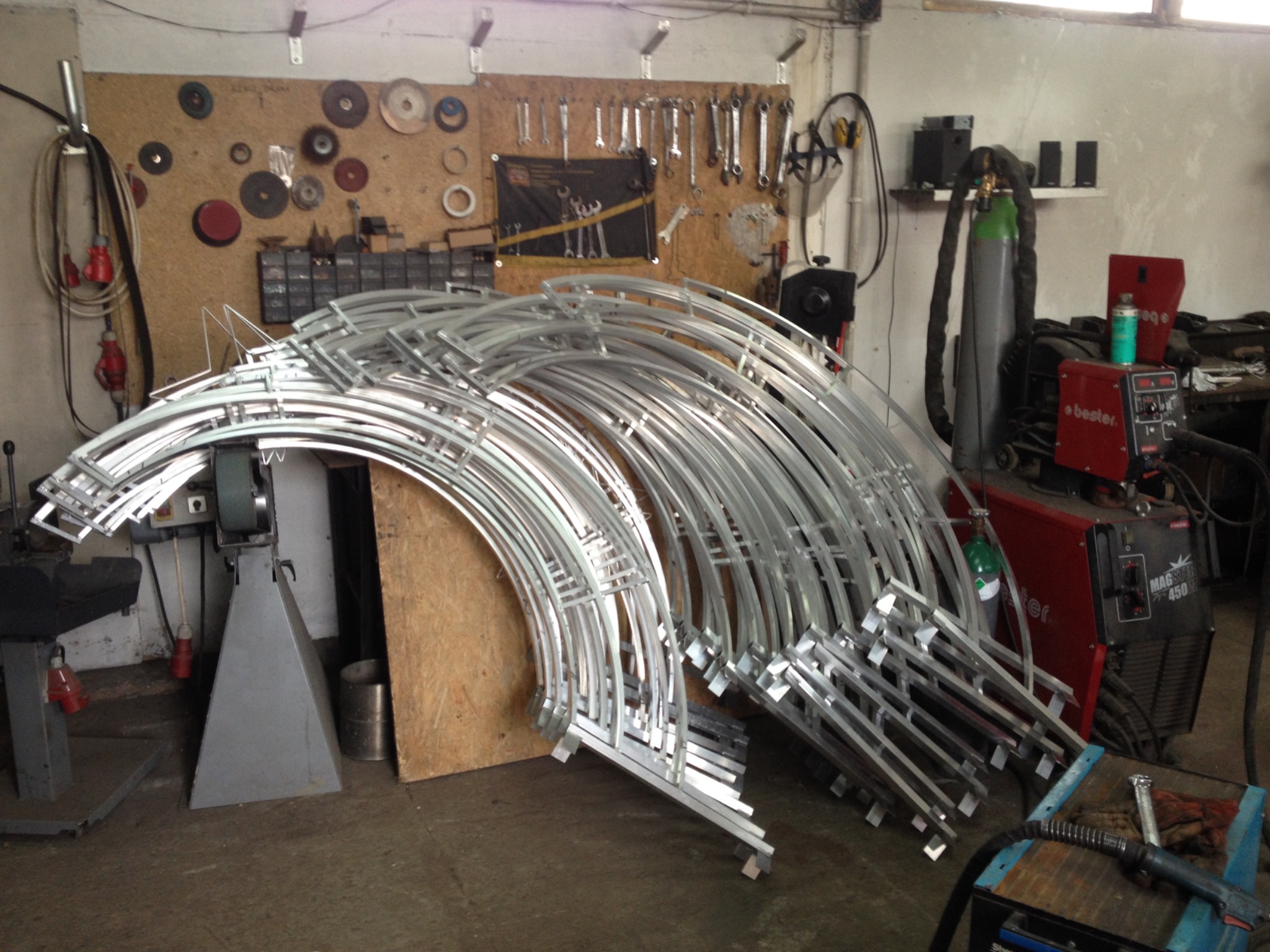 Stos wygiętych, metalowych elementów konstrukcyjnych w warsztacie, w tle narzędzia i spawarka.