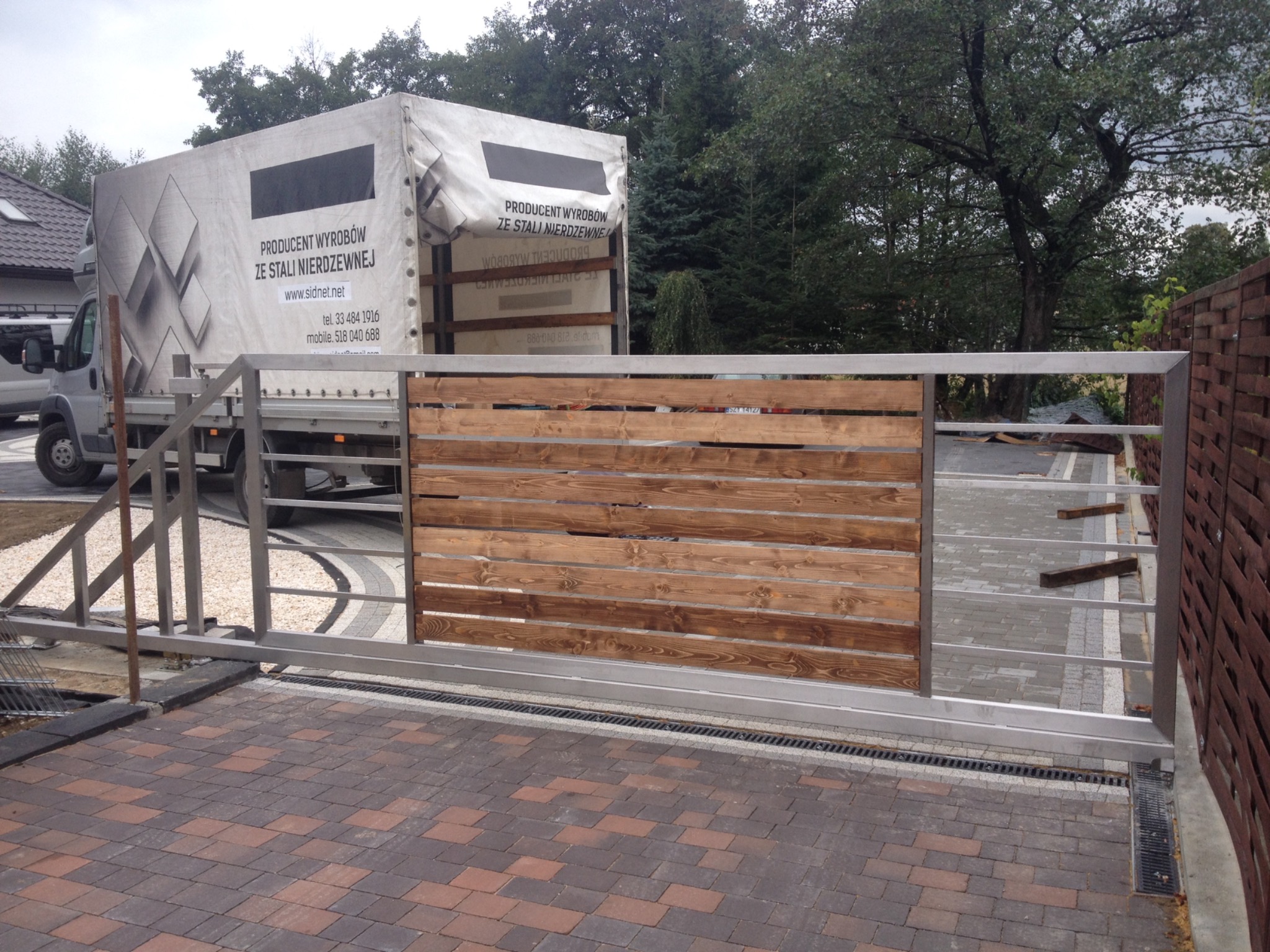 Przesuwna brama wjazdowa z drewnianym wypełnieniem i metalową ramą, widoczna na tle brukowanej posesji i zaparkowanego samochodu dostawczego z napisem 'Producent wyrobów ze stali nierdzewnej'.